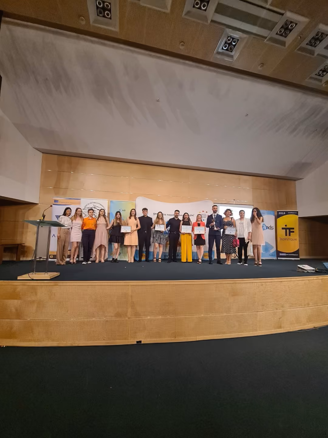 Tineri pe o scenă, ținând diplome, în fața bannerelor partenerilor