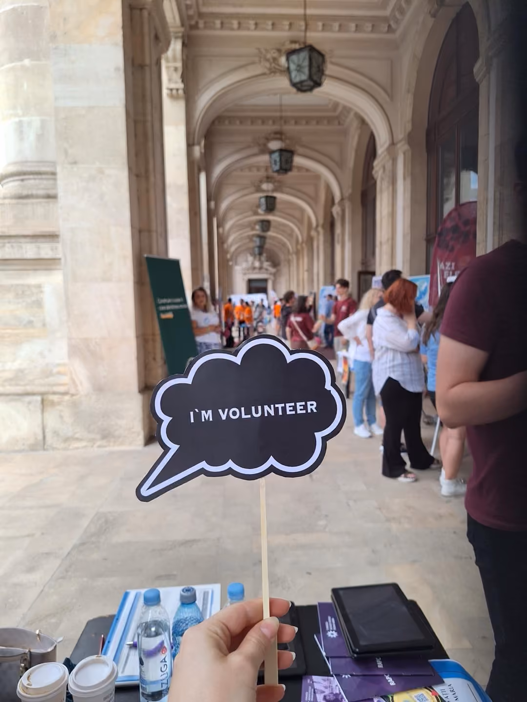 O mână ține un panou în formă de balon de dialog cu mesajul „I'm volunteer”, în fața standurilor târgului