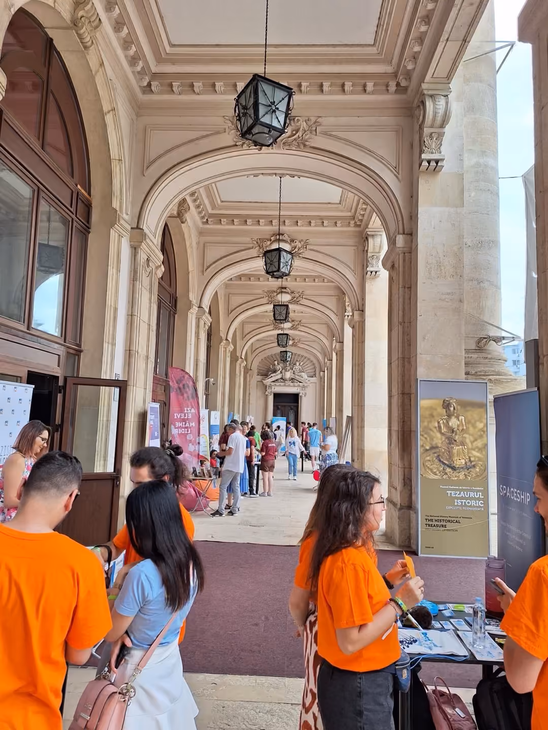 Voluntari în tricouri portocalii la un stand, în galeria cu arcade a Muzeului Național de Istorie