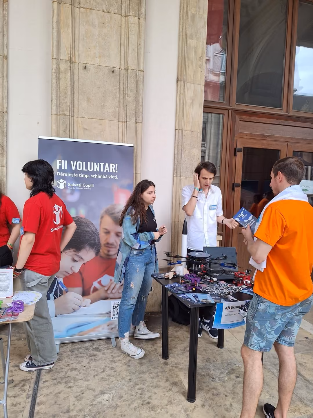Voluntari discutând la standul Salvați Copiii, lângă un banner „Fii voluntar!”